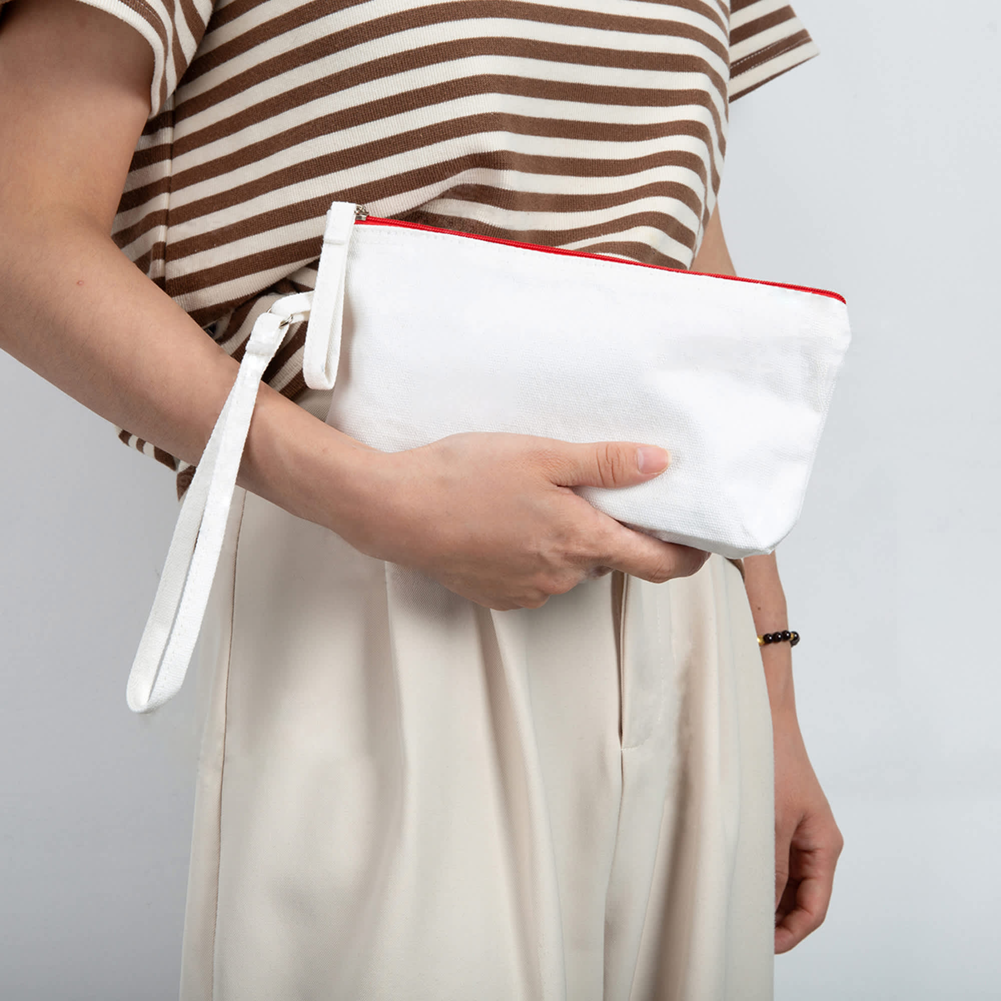 A white canvas cosmetic bag with a red zipper, held by a person wearing a striped top and light trousers.