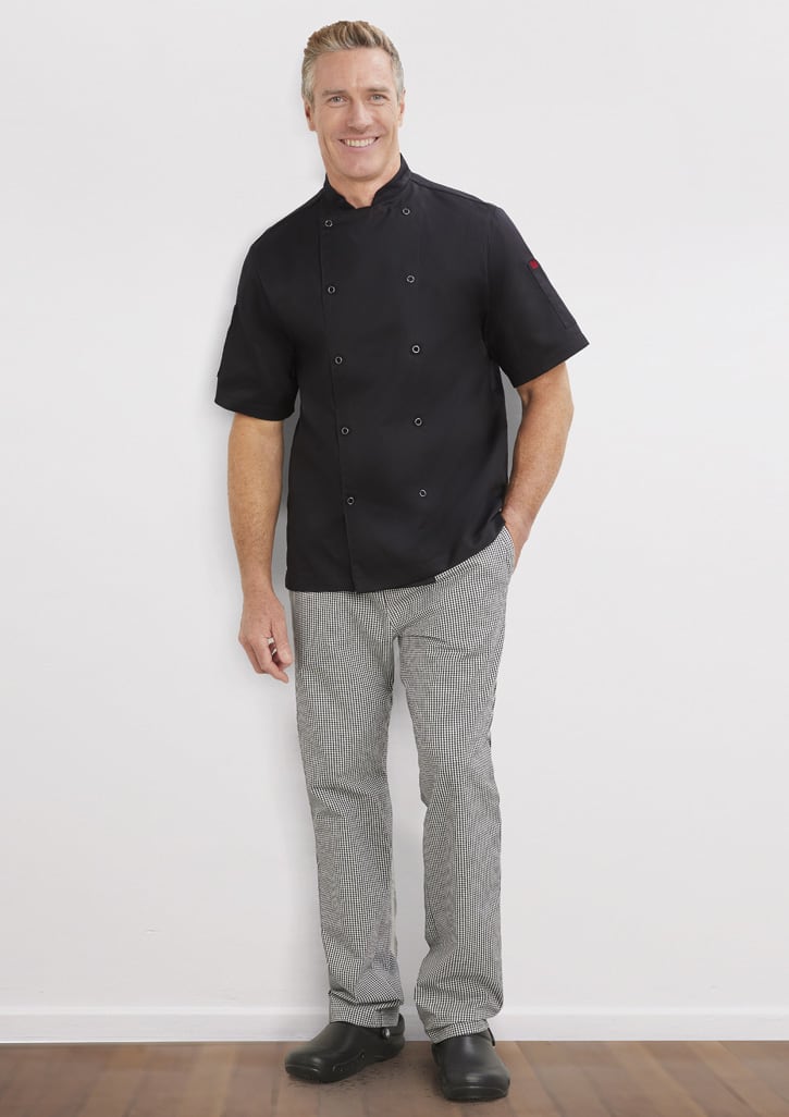 A man stands smiling, wearing a black short-sleeve chef's jacket and grey striped pants with a logo on the jacket.