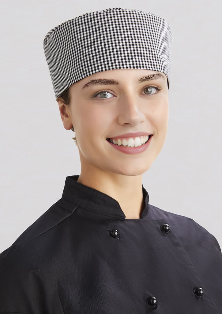 A chef cap in a black and white checkered pattern, worn by a smiling individual in a black chef's uniform.