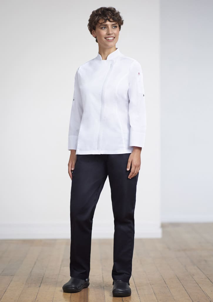 A woman stands wearing a white uniform top and navy scrub bottoms, paired with black shoes, in a bright indoor setting.