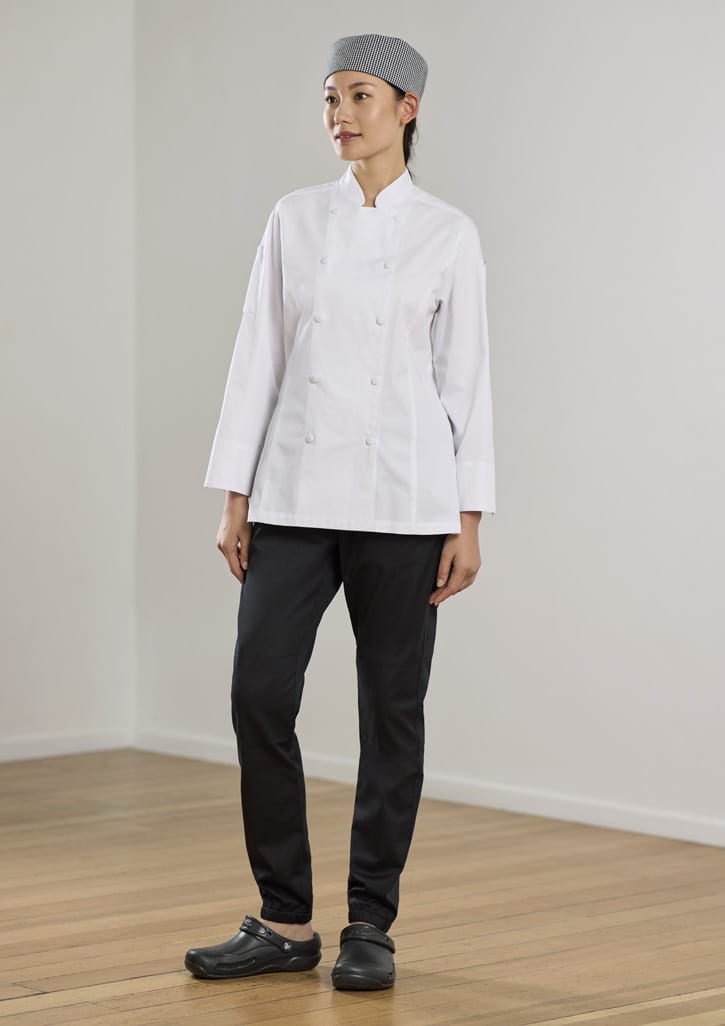 A woman in a white chef jacket and black jogger pants stands against a neutral background. She wears a grey checked head covering and clogs.