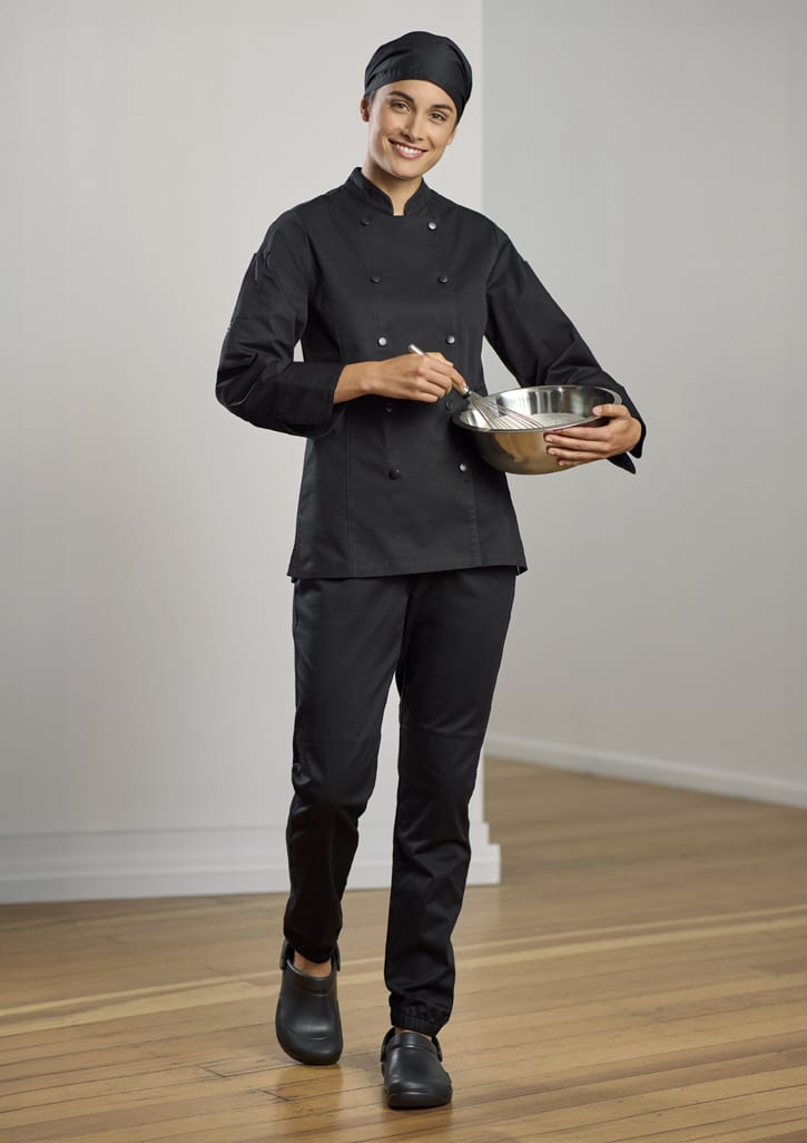 A woman in a black chef uniform and matching jogger pants holds a mixing bowl, smiling in a bright kitchen setting.