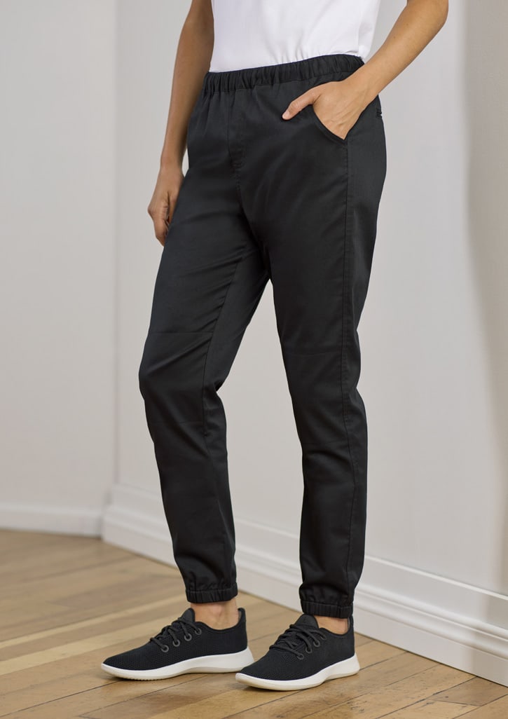 Black jogger pants with an elastic waistband and pockets, styled for women's chefwear. The model pairs them with black sneakers.