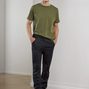A man stands wearing olive green t-shirt and black jogger pants, with hands in pockets. The setting is minimalistic.