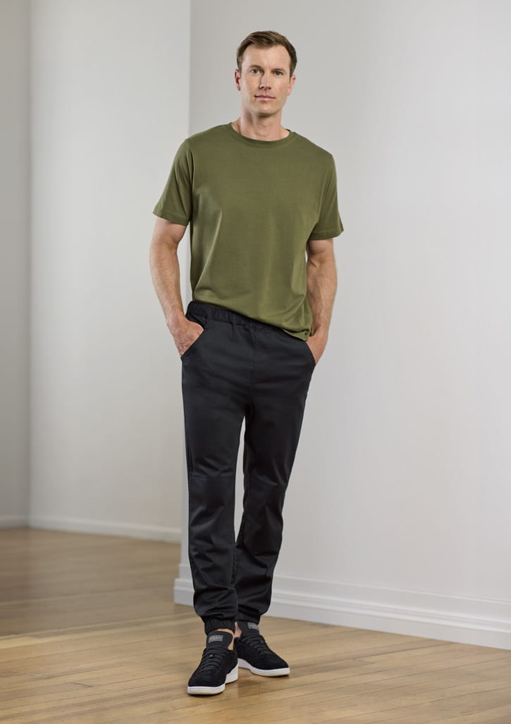 A man stands wearing olive green t-shirt and black jogger pants, with hands in pockets. The setting is minimalistic.