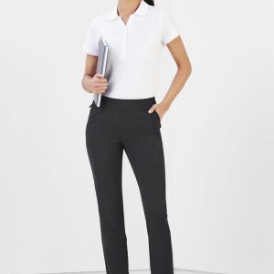 A woman stands wearing black Jane Stretch Pants with a white polo shirt, holding a notebook in a minimalist setting.
