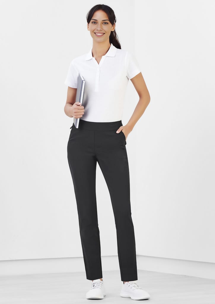 A woman stands wearing black Jane Stretch Pants with a white polo shirt, holding a notebook in a minimalist setting.