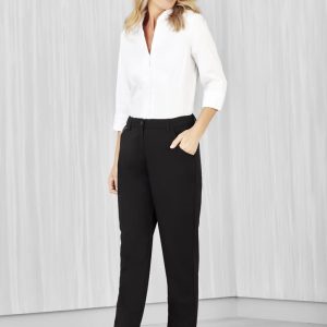 A woman stands wearing black straight leg pants and a white blouse, smiling. The pants have pockets and a logo.