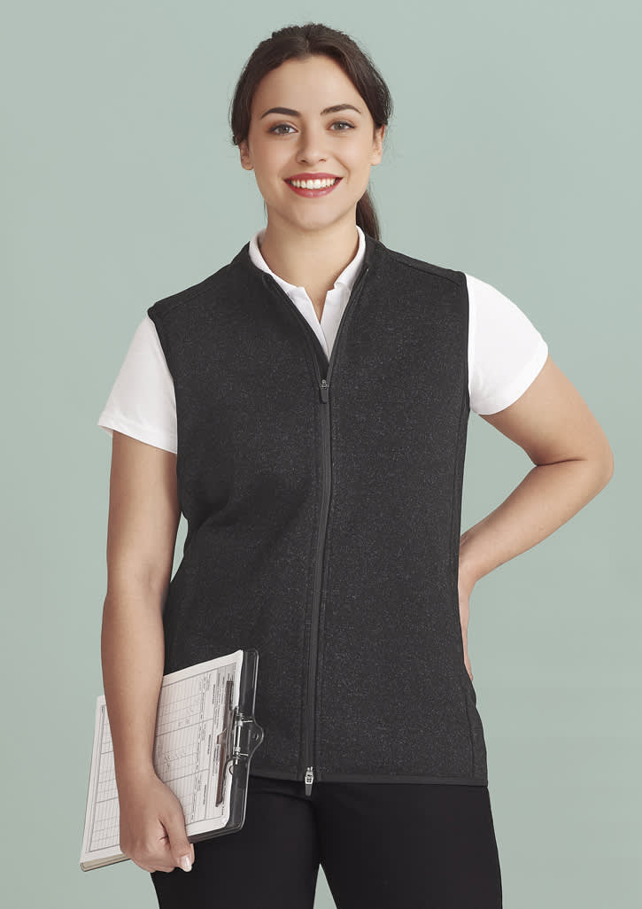 A woman wearing a black vest with a zip front, holding a clipboard, against a light green background.