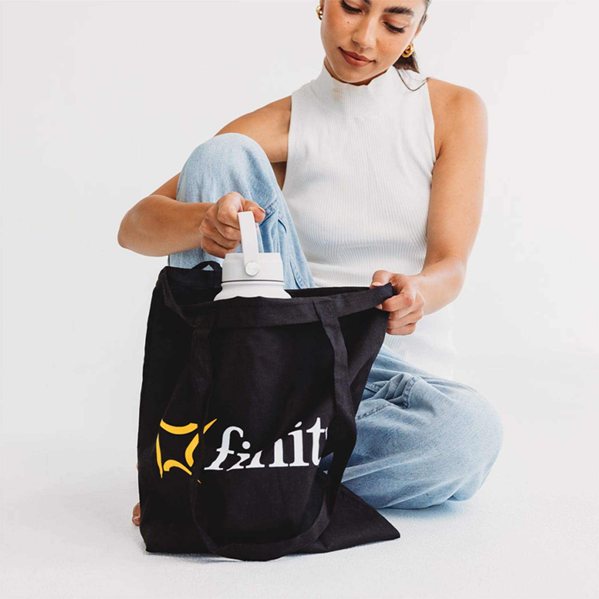 A black tote bag with a logo, being held by a person, while they reach inside for a drink bottle.