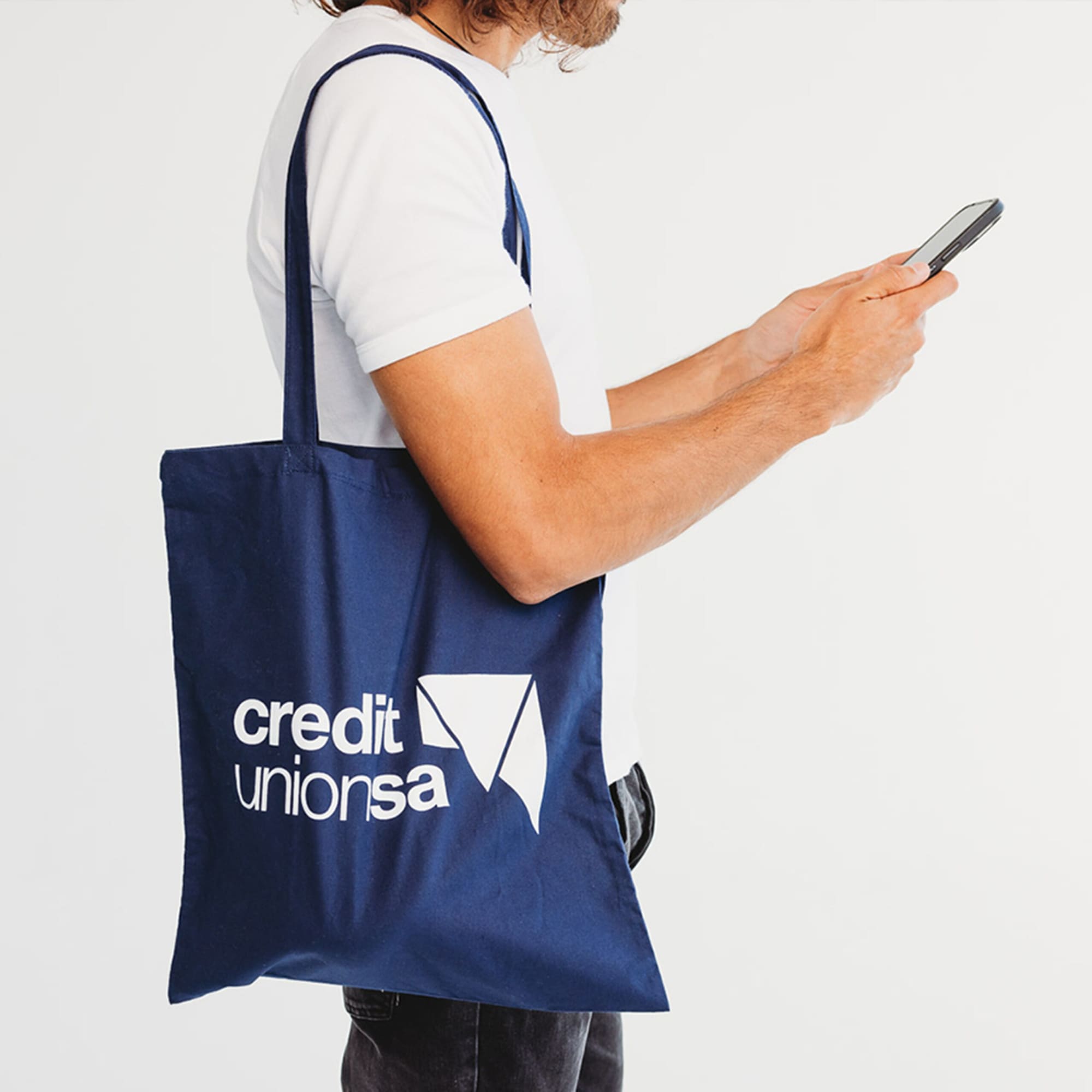 A navy blue tote bag with a logo sits over the shoulder of a person holding a smartphone.
