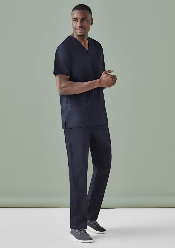 A man is wearing navy unisex scrub pants and a matching scrub top, standing against a plain green background.