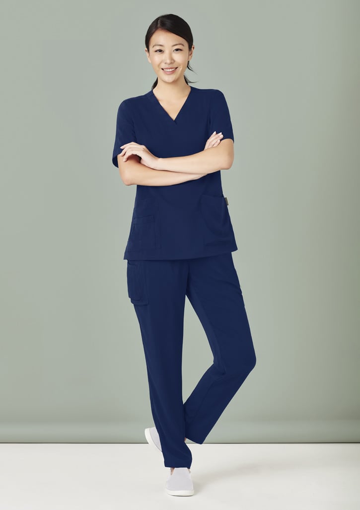 Navy blue women's scrub top and slim leg pant set, featuring pockets and a branding logo. Model is smiling.