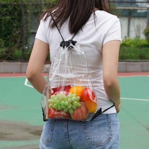 A transparent drawstring backpack filled with assorted fruits, held by a person wearing a white shirt and denim shorts.