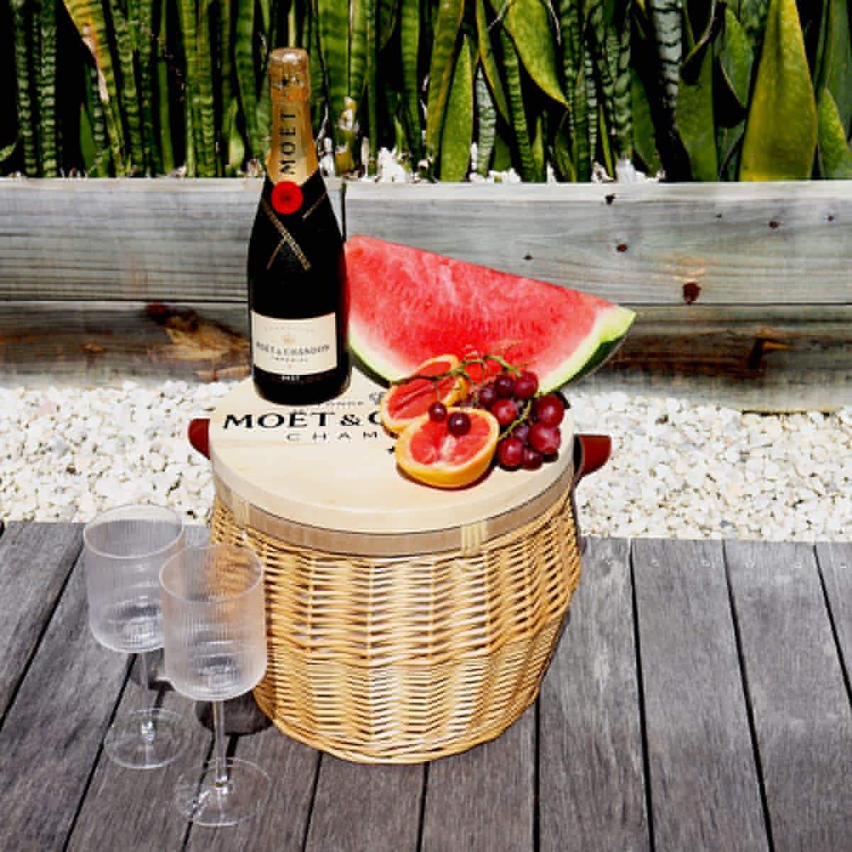 A wicker cooler basket with a wooden lid, a bottle of champagne, watermelon, grapes, and two glasses.