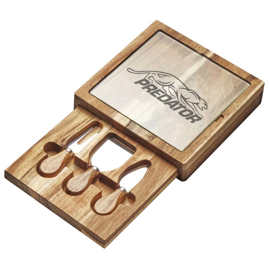 A cheese board set featuring a glass top and wooden base, including three stainless steel tools and a logo.