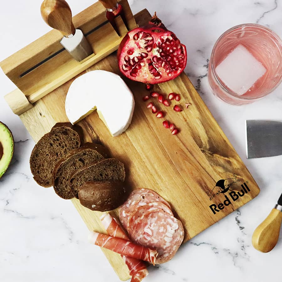 A wooden cheeseboard with a cheese wedge, sliced meats, pomegranate, and bread, featuring a logo.