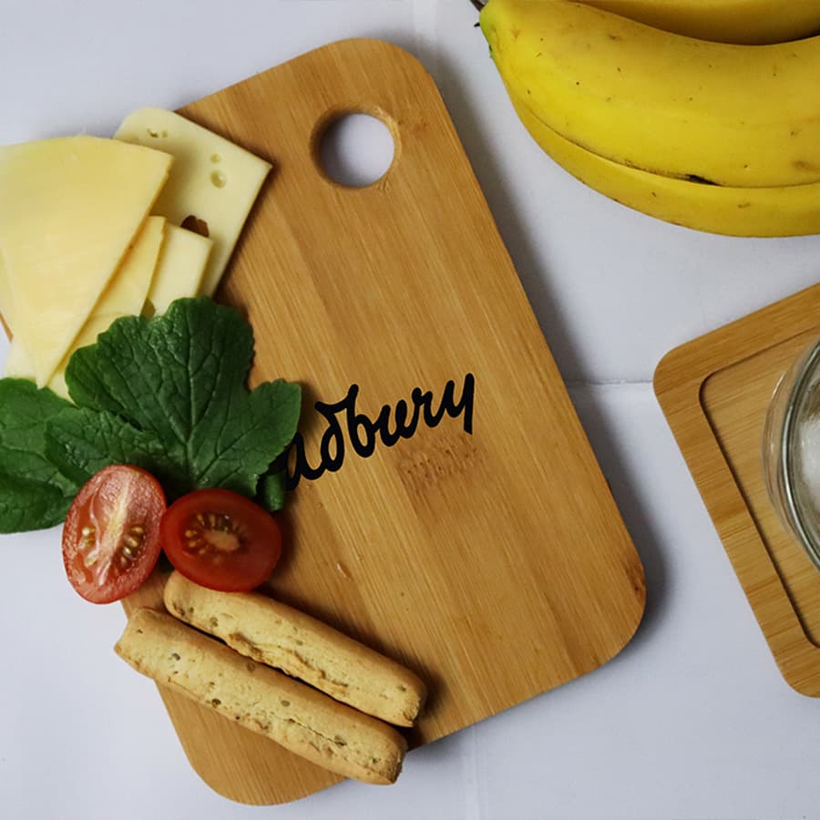 A small bamboo cutting board with cheese, tomatoes, mint leaves, and snacks, featuring a logo, beside ripe bananas.