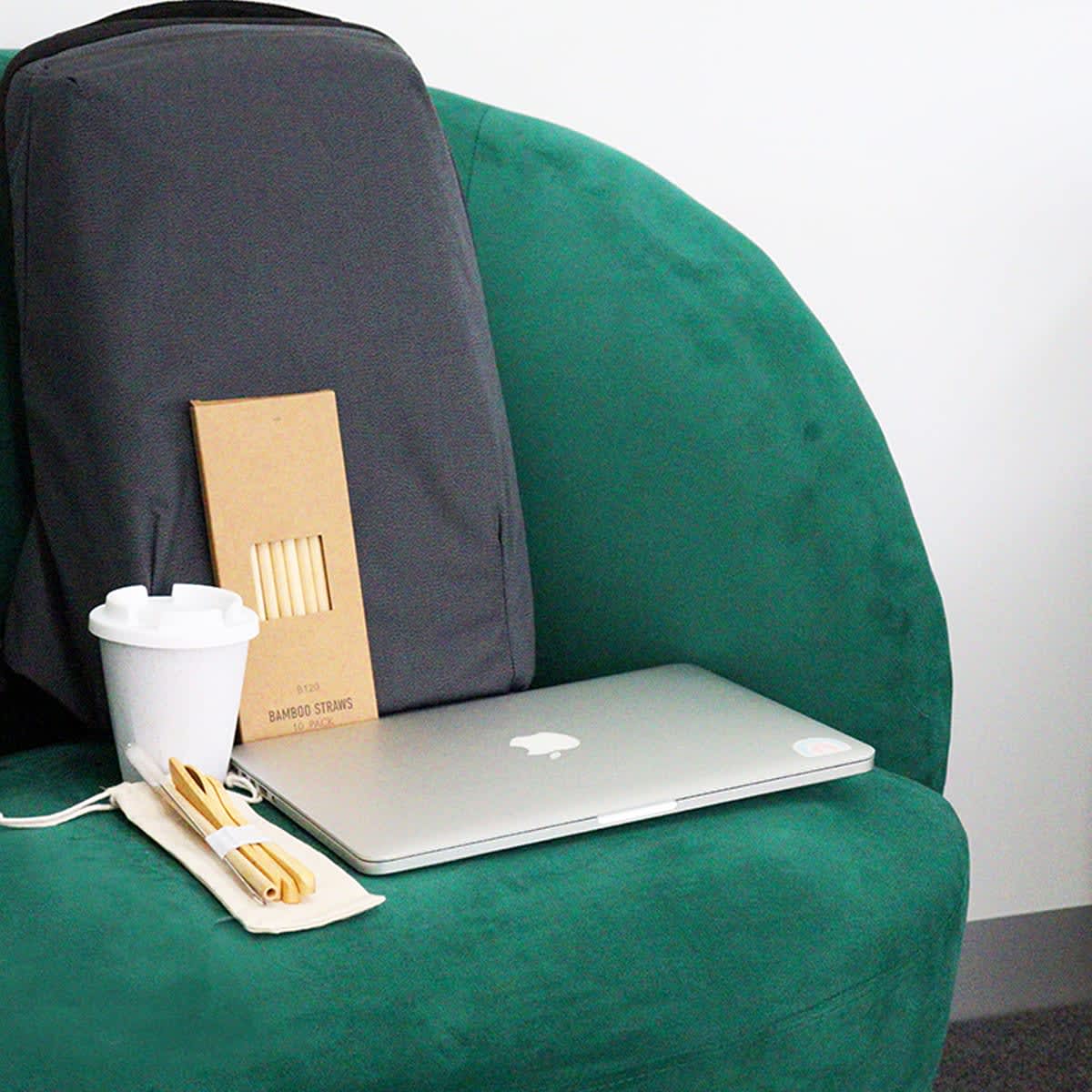 A black Nova Computer Backpack rests on a green chair, alongside a laptop, reusable cup, and bamboo straws.