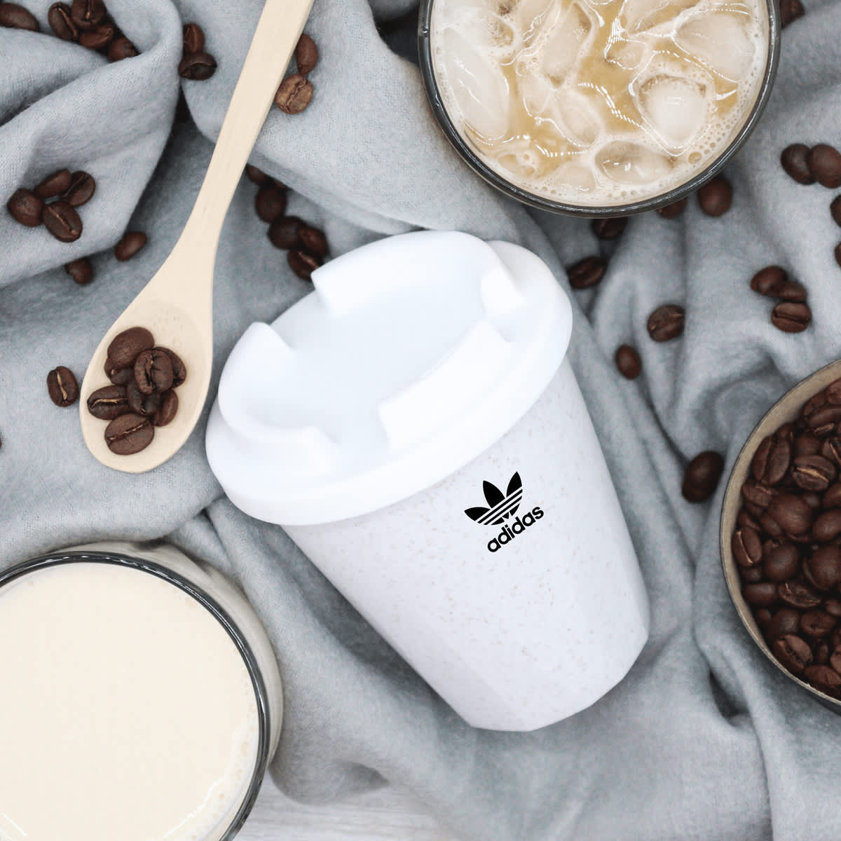 A white 350mL cup with a logo, surrounded by coffee beans and beverages on a grey fabric backdrop.