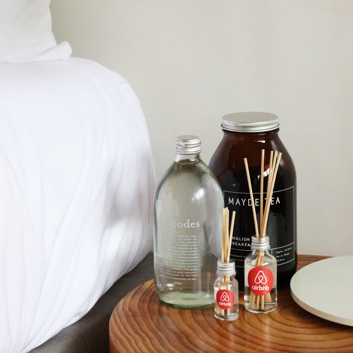A glass diffuser bottle with reed sticks alongside a larger tea container, set on a wooden table near a bed.