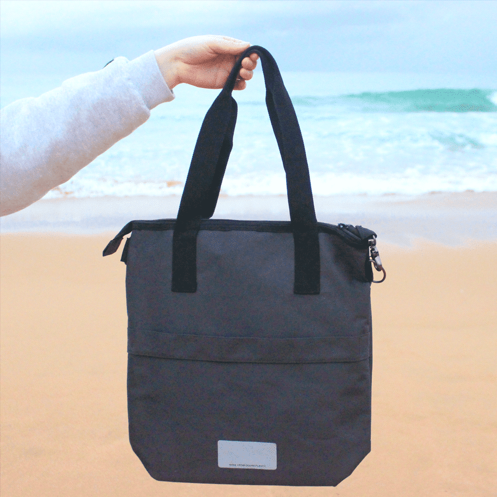 A black laptop tote bag made from recycled material, featuring two handles and a zipper closure, being held by a person.