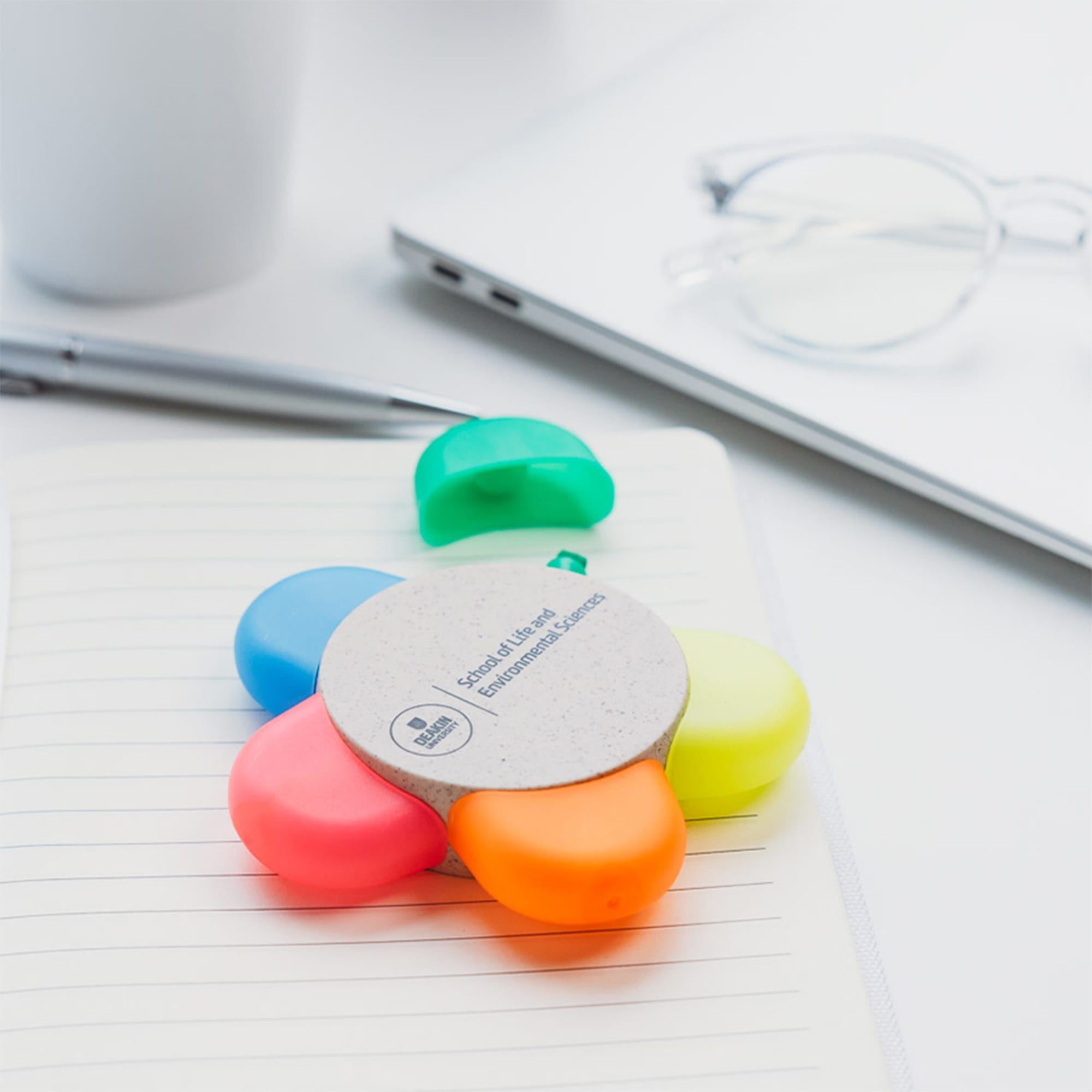A flower-shaped set of highlighters in pink, orange, yellow, green, and blue, on a notebook with a pen and glasses nearby.