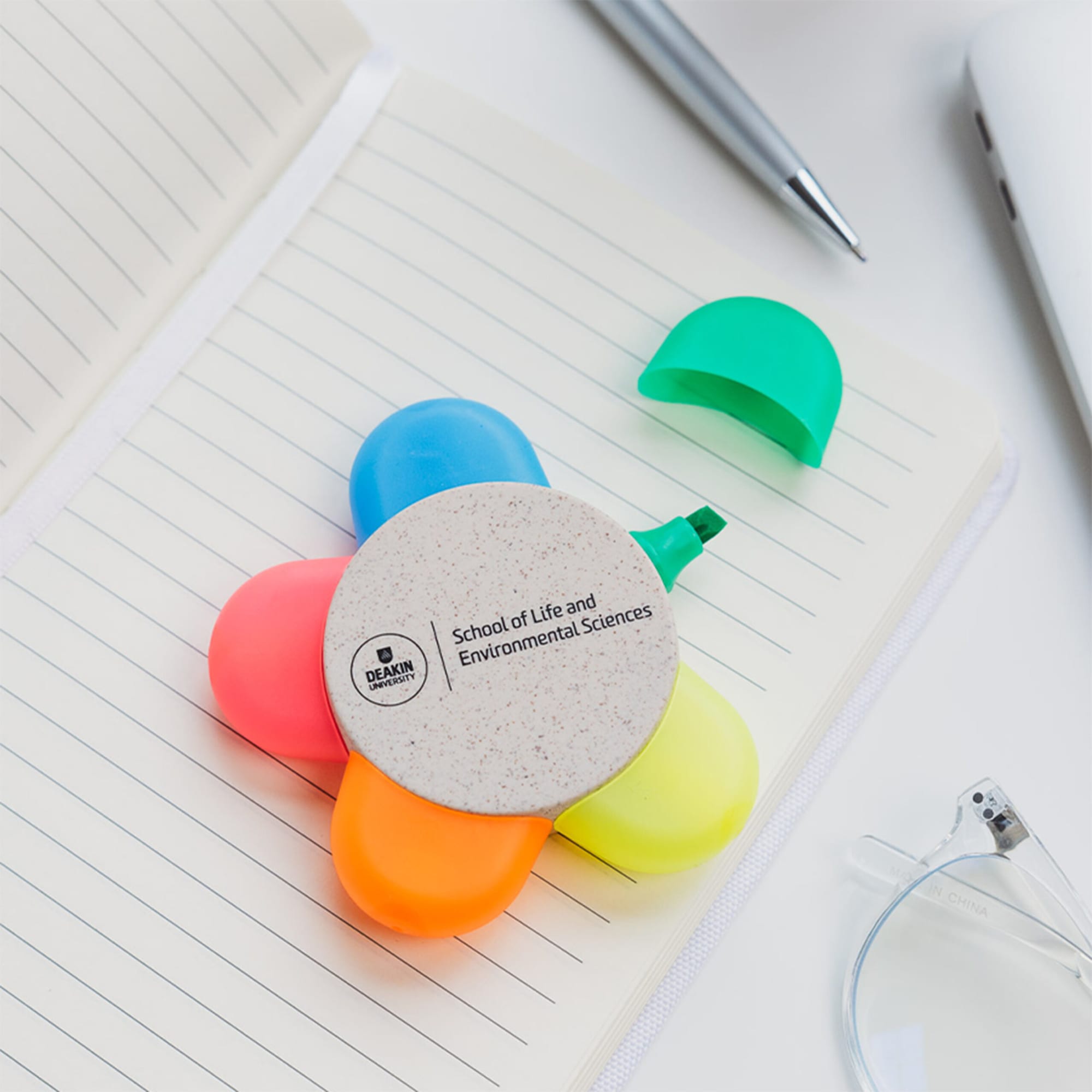A flower-shaped set of highlighters in pink, orange, green, and yellow on a notebook. It features a logo on the centre.