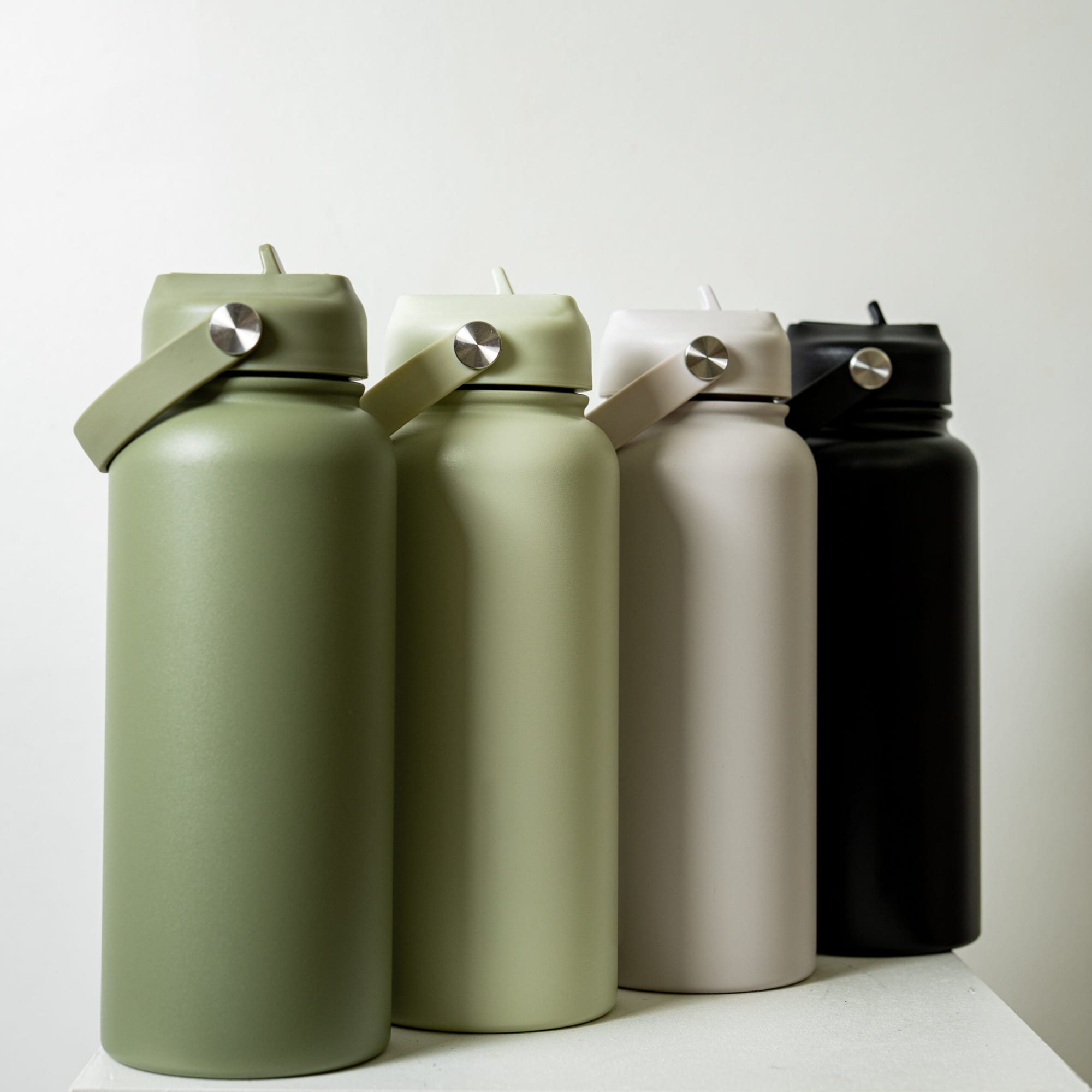 A drink bottle featuring a matte finish, available in olive green, cream, and black, with a strap and a logo.