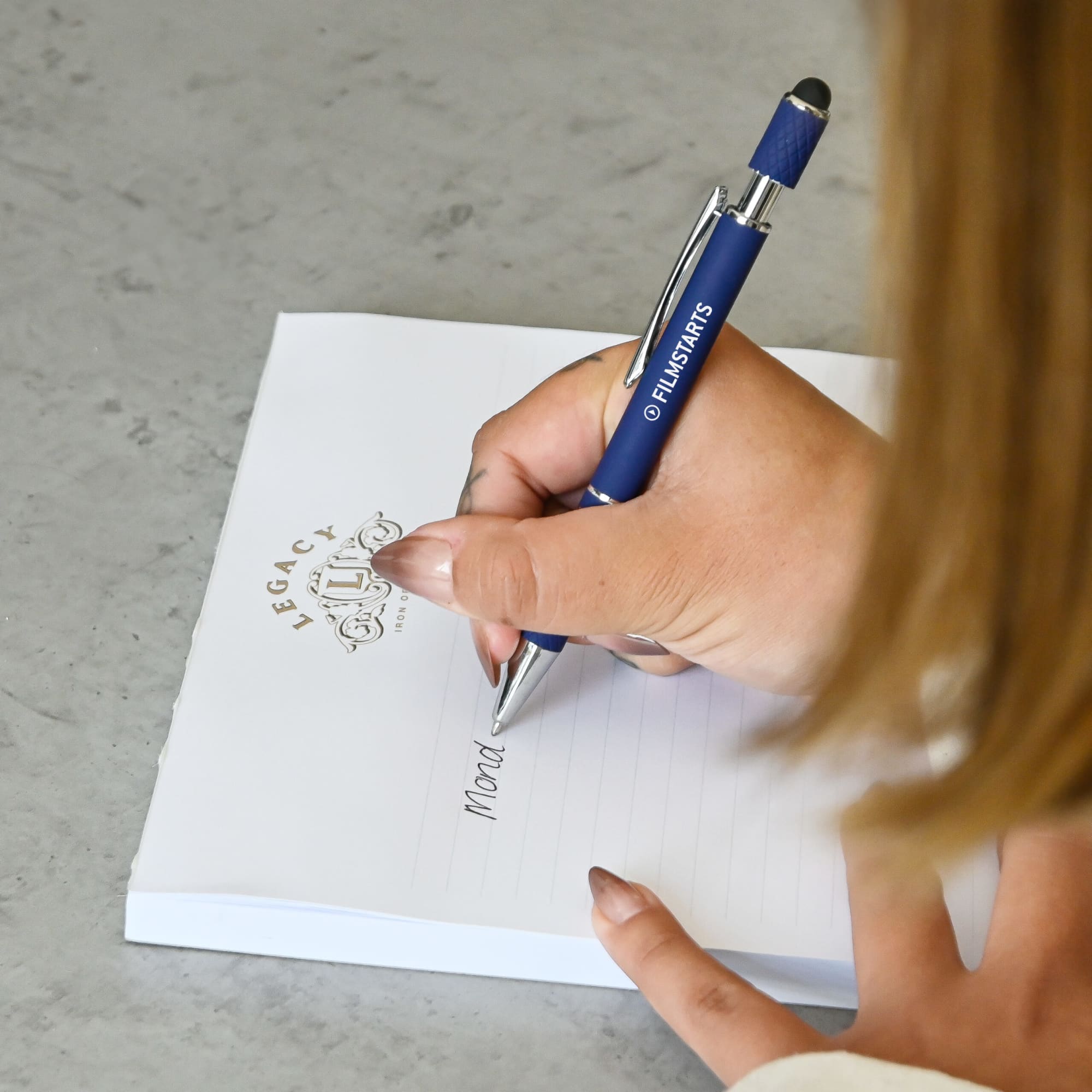 A blue metal pen with a logo is being used to write on a notepad with a decorative header.