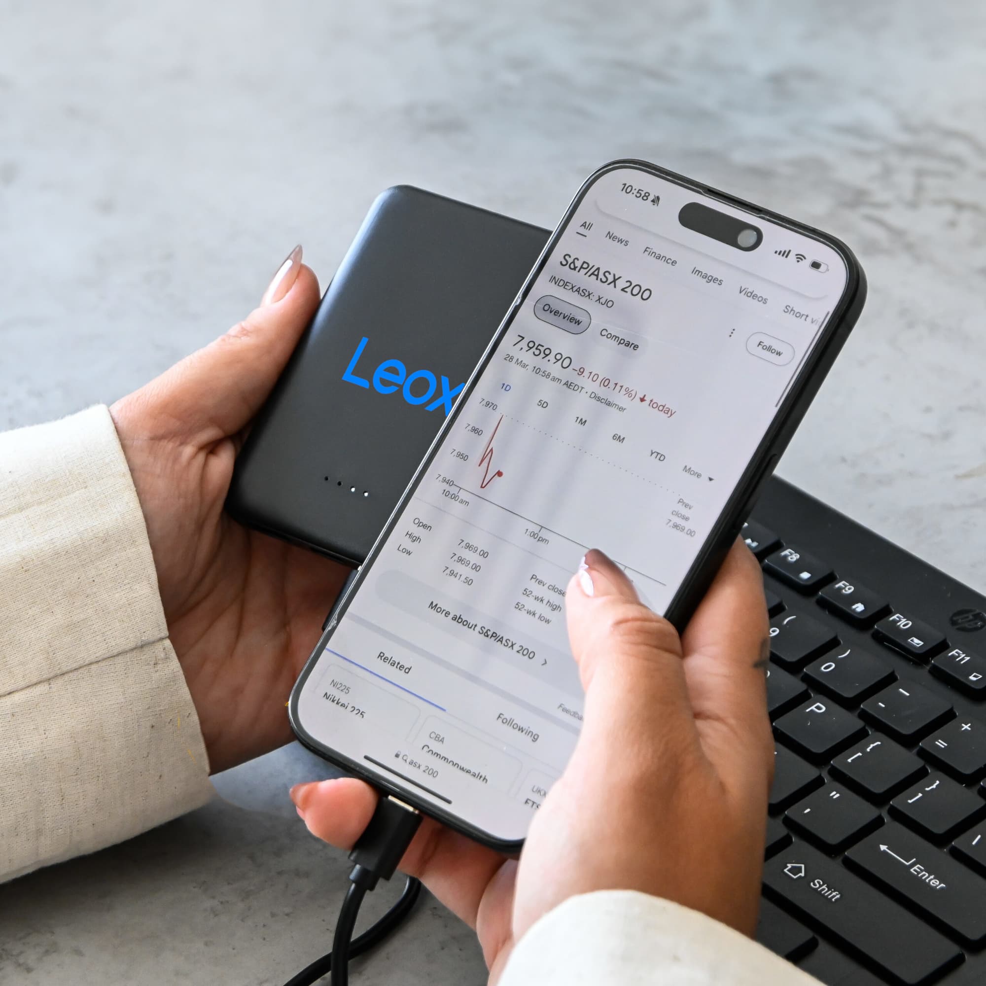 A black power bank is held in one hand while the other hand holds a smartphone displaying financial data, with a keyboard in the background.