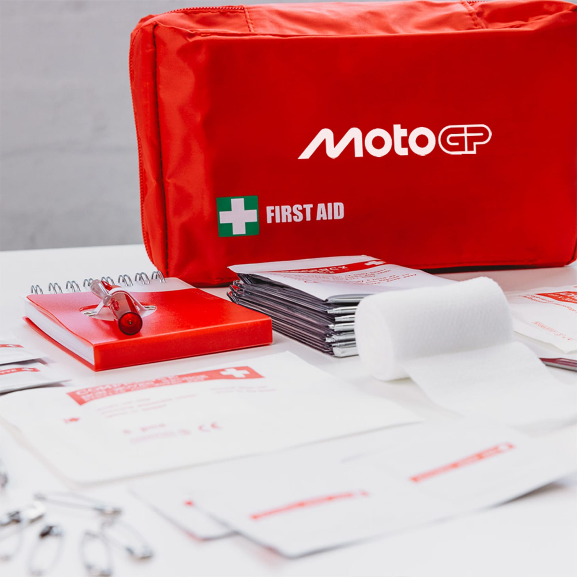 A red first aid kit with various contents including bandages, a notebook, scissors, and safety pins, featuring a logo.