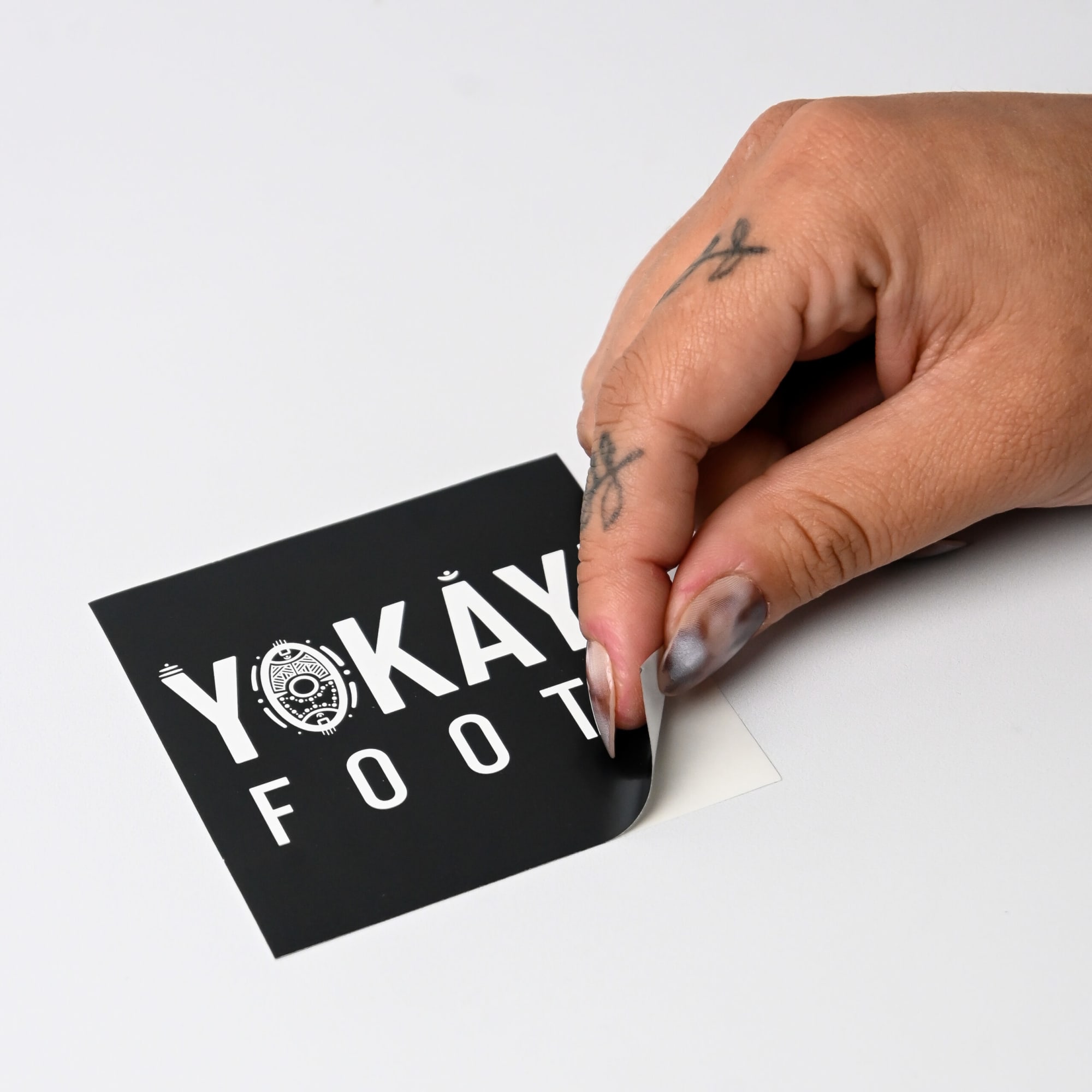 A hand with tattoos is peeling a black gloss paper sticker featuring bold white text and a circular design.