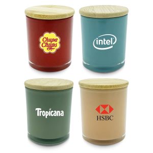 Four large candles in various colours: red, green, blue, and beige, each with a wooden lid and branded logos.