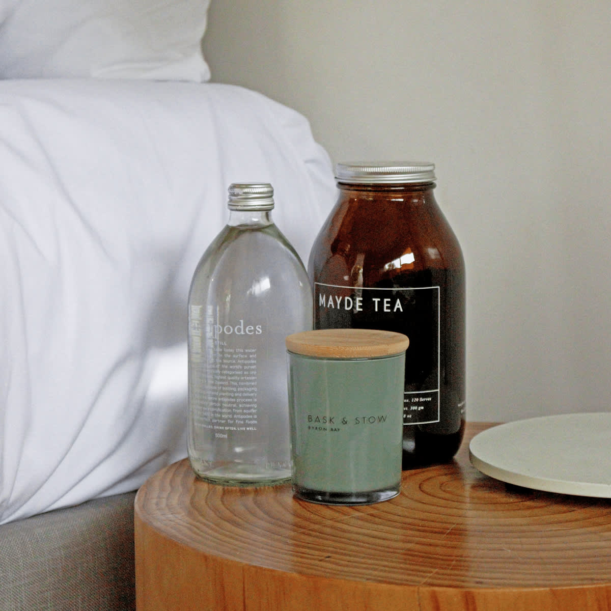A medium green candle with a wooden lid, alongside a glass bottle and a jar of tea on a wooden table.