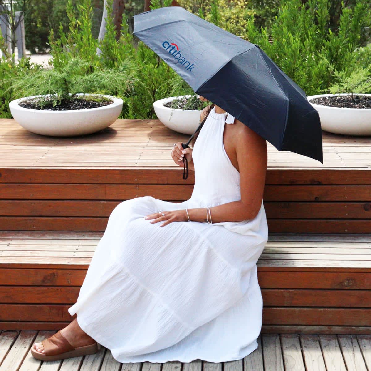 A black umbrella with a logo, held by a person in a white dress, sitting on wooden steps with greenery in the background.