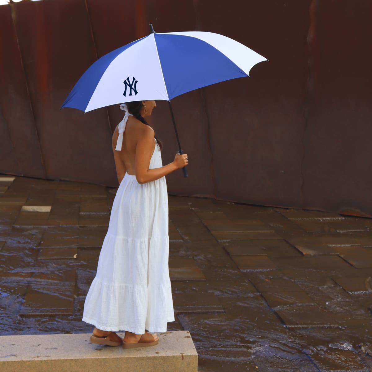 A blue and white umbrella with a logo, held by a person in a white dress, standing near water.
