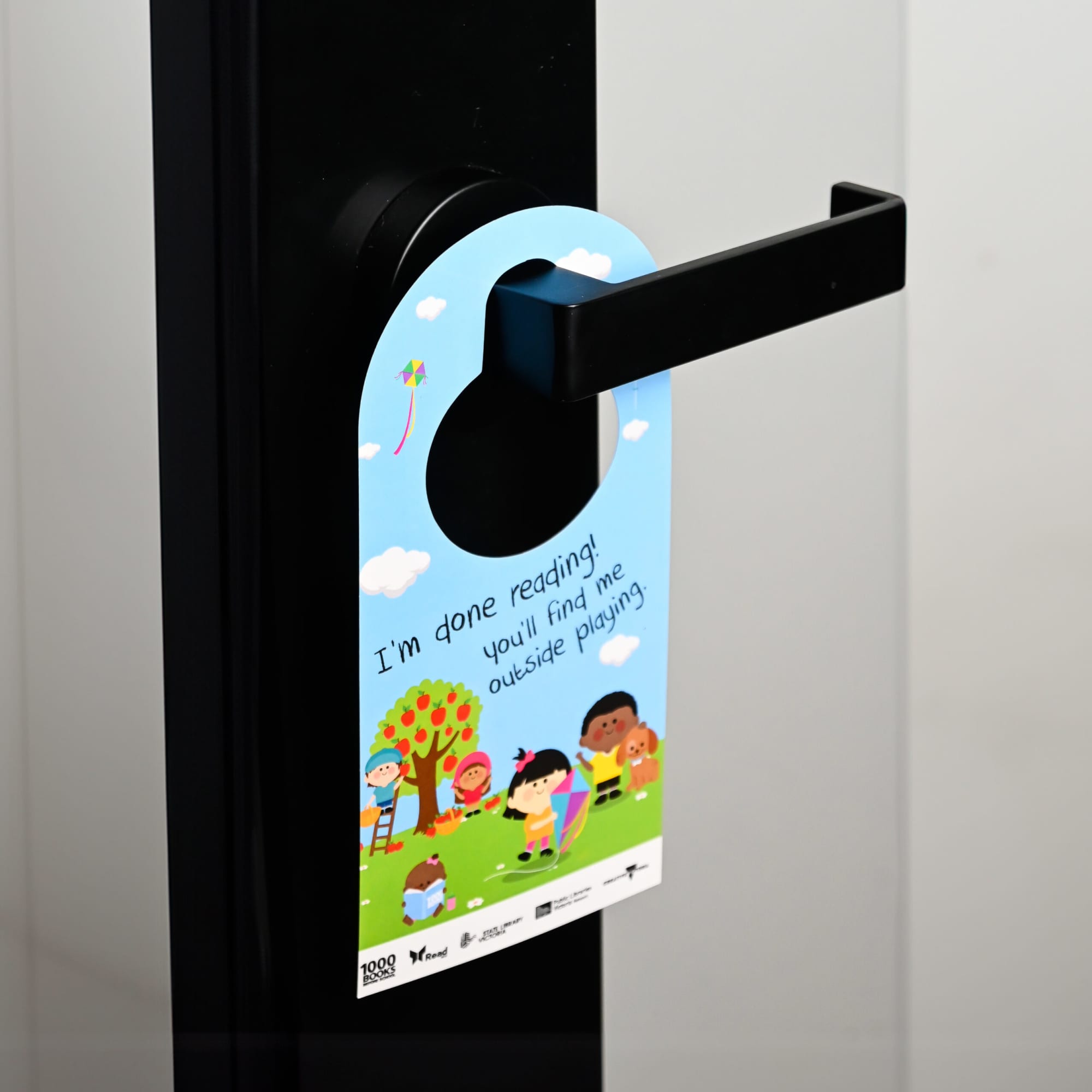 A colourful door hanger featuring children playing outdoors, attached to a black door handle.