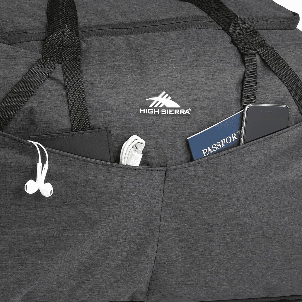 A person opens a charcoal grey duffel bag, revealing a cozy interior with a blanket inside. The bag has a logo.