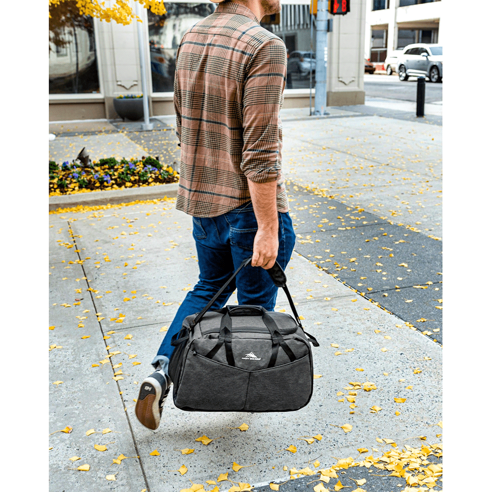 A dark grey duffel bag with a logo, placed on a table beside a potted plant and woven basket.