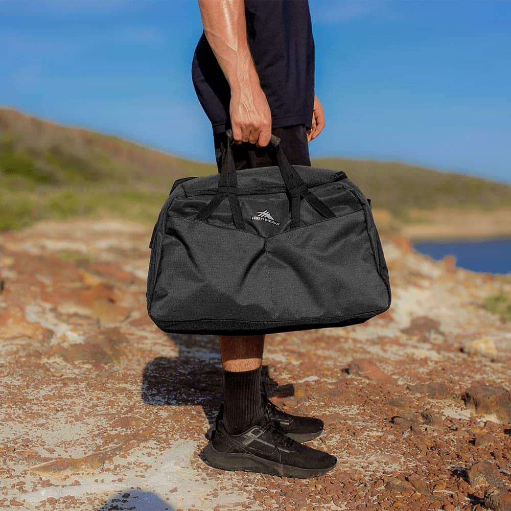 A grey duffel bag with multiple pockets and a logo, resting on a table next to a person sitting on a couch.