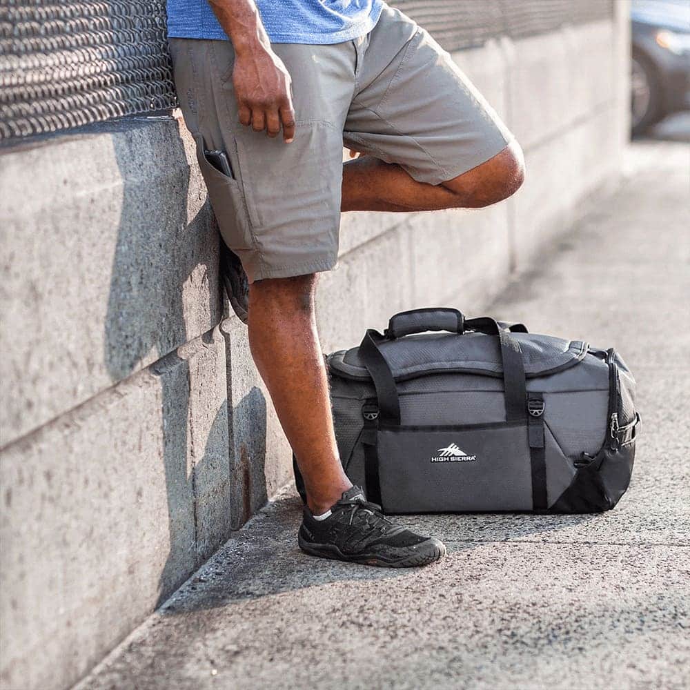 A duffel backpack in grey with black accents, resting on the ground beside a person in shorts and sneakers.