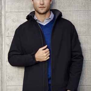 A black soft shell jacket is worn by a man against a concrete wall. The jacket has a hood and a logo on the sleeve.