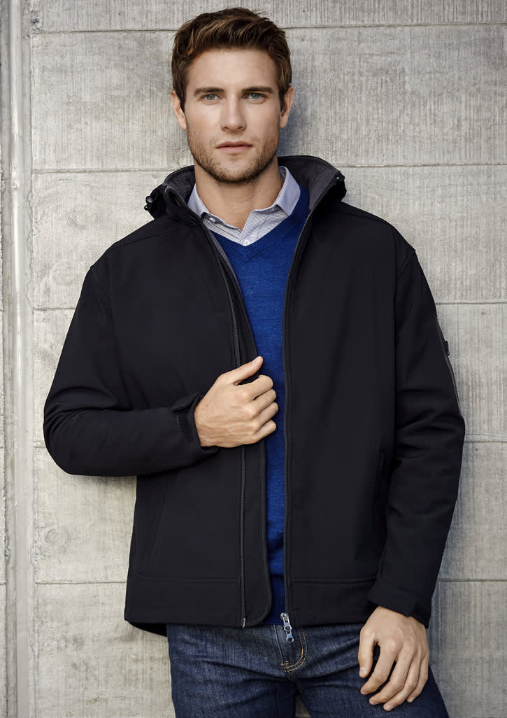 A black soft shell jacket is worn by a man against a concrete wall. The jacket has a hood and a logo on the sleeve.