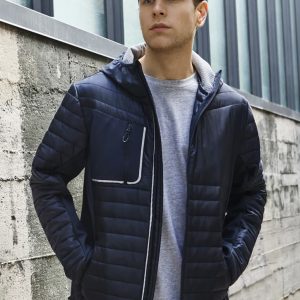 A unisex navy soft shell jacket with a zippered pocket, worn by a model against an industrial backdrop.