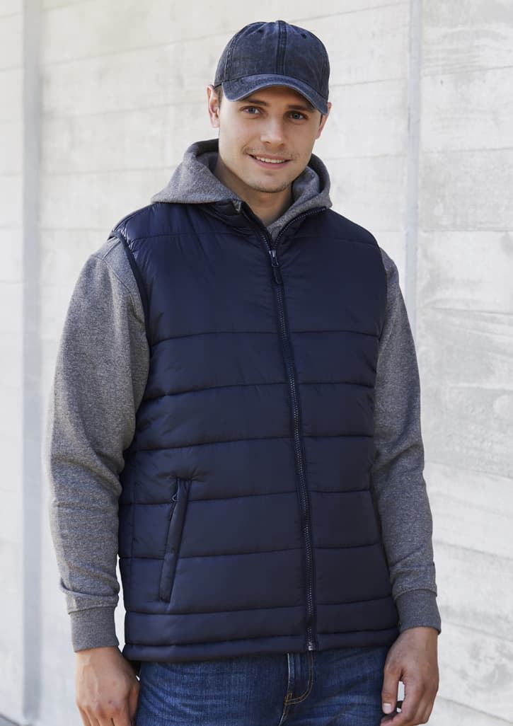 A man wearing a navy blue puffer vest over a grey hoodie, combined with denim jeans and a cap. The vest has a front zipper and pockets.