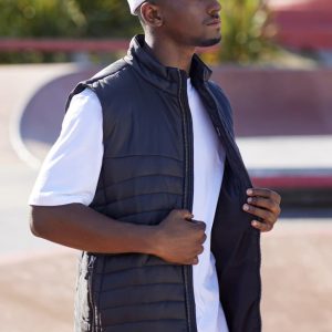 A man wears a black men's expedition vest, standing outdoors, with a white cap and t-shirt. The vest is unzipped.
