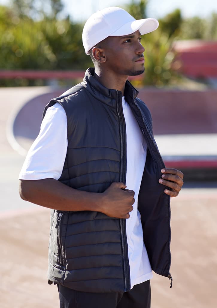 A man wears a black men's expedition vest, standing outdoors, with a white cap and t-shirt. The vest is unzipped.