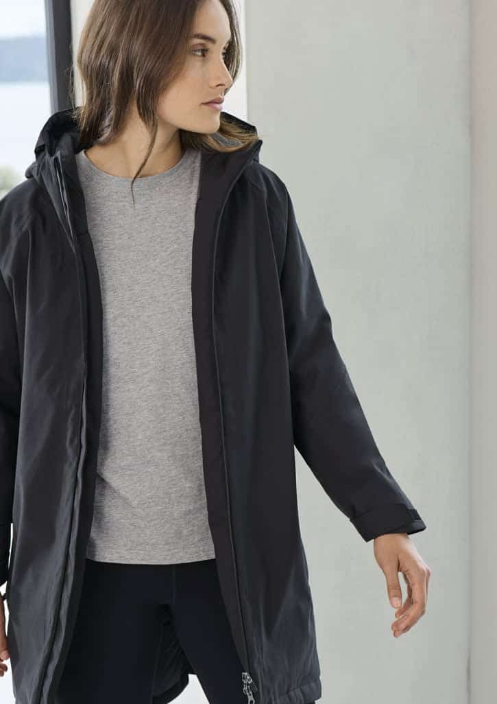 A unisex black rain jacket with a hood, worn over a grey top, showcased in a light-filled setting.
