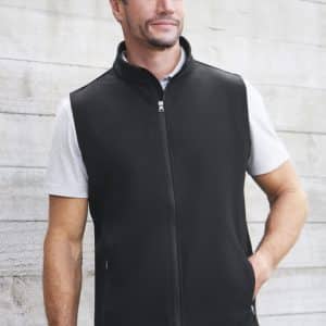 A man models a black vest with a zip front and side pockets against a rustic background. The vest has a logo.